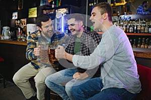Three young men in casual clothes are smiling and clanging glasses of beer together while sitting at bar counter in pub