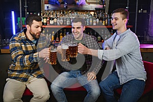 Three young men in casual clothes are smiling and clanging glasses of beer together while sitting at bar counter in pub