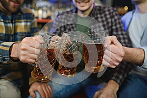 Three young men in casual clothes are smiling and clanging glasses of beer together while sitting at bar counter in pub