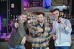 Three young men in casual clothes are smiling and clanging glasses of beer together while sitting at bar counter in pub