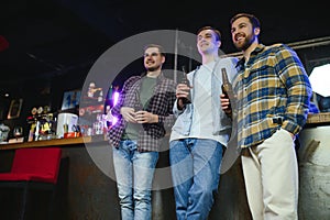 Three young men in casual clothes are smiling, holding bottles of beer while standing near bar counter in pub