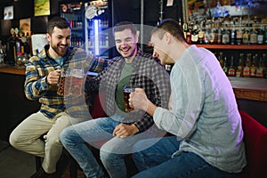 Three young men in casual clothes are smiling, holding bottles of beer while standing near bar counter in pub
