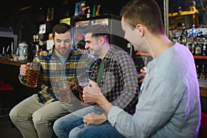 Three young men in casual clothes are smiling, holding bottles of beer while standing near bar counter in pub