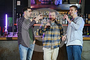 Three young men in casual clothes are smiling, holding bottles of beer while standing near bar counter in pub