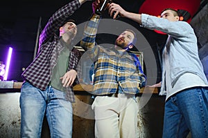 Three young men in casual clothes are smiling, holding bottles of beer while standing near bar counter in pub