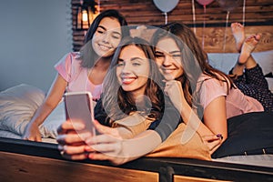 Three young models taking selfie. They pose and smile. Gils are in room. One of them hold phone and take picture on it