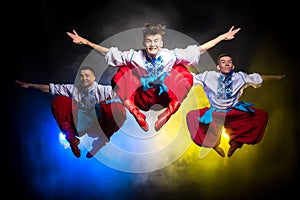 Three young people in the Ukrainian national costumes dance and jump on a dark background with smoke