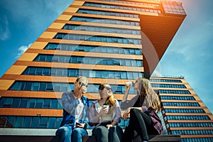 Three young pretty girlfriends sitting on bench on city street and talking. Women drink coffee from disposable glasses