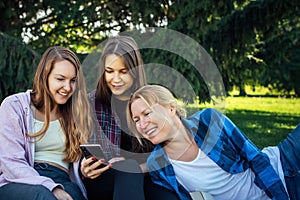 Three young pretty women on the lawn in park look at smartphone and laugh. Female students relax in the park sitting on green