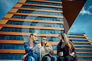 Three young pretty women sit on a bench on a city street, drinking coffee from disposable glasses, talking and laughing. Friends