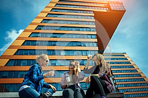 Three young pretty women sit on a bench on a city street, drinking coffee from disposable glasses, talking and laughing. Friends