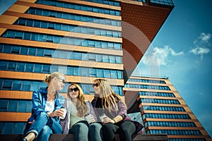 Three young pretty women sit on a bench on a city street, drinking coffee from disposable glasses, talking and laughing. Friends