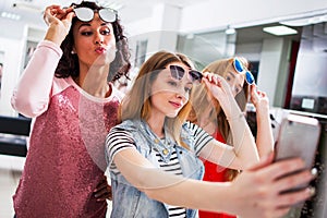 Three young stylish girlfriends raising fashionable sunglasses while taking selfie with smartphone in shopping mall