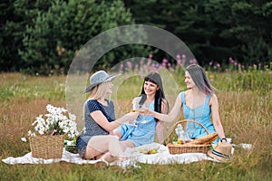 Three young women, blond, brunette and with dyed hair in blue dresses, and hats, sit on plaid and drink wine from