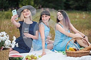 Three young women, in blue dresses and hats sit on a plaid and take pictures on a smartphone. Outdoor picnic on the