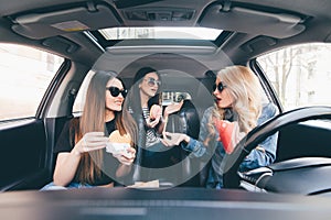 Three young women drive a car, speaking each other and eating a fast food in a car in traffic jam