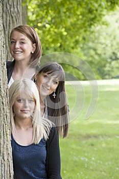Three young women looking from behind a tree