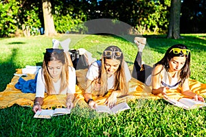 Three young women students spending time lying on meadow in city park at sunset or dawn reading book to learn university school