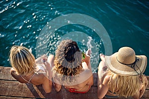 Three young women in swimsuits sitting by beautiful azure blue water, with drinks. Summertime vacation with girlfriends. Holiday,