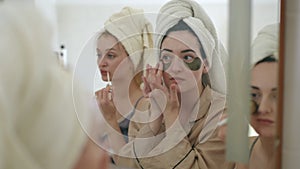 Three young women with towels on their heads do morning beauty treatments while standing in front of a mirror in the