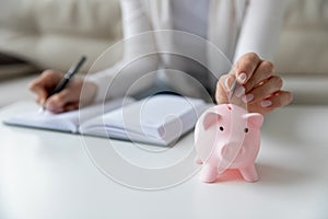 Thrifty woman writing daily expenses put coin into piggy bank