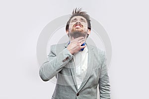 Throat pain. Portrait of sick handsome bearded man in casual grey suit and blue bow tie standing and holding his painful throat