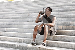 Thursty Black Male Athlete Drinking Water, Resting On Urban Stairs After Training Outdoors