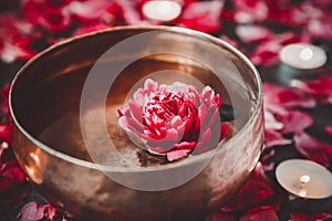 Tibetan singing bowl with floating inside in water red peony flower. Burning candles and petals on the black stone background.