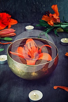 Tibetan singing bowl with floating lily inside. Burning candles, lily flowers and petals on the black wooden background. Meditatio