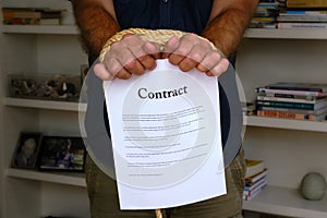 Tied hands holding contract paper in front of bookcase. Bound man hands with paper contract, breach of contract. Unfair