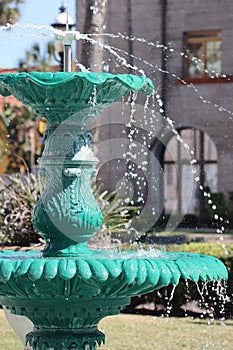 Tiered verdigris metal fountain with spraying water and gray stone building in background photo