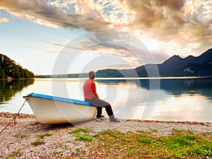Tiired man in red shirt sit on old fishing paddle boat at mountains lake coast.
