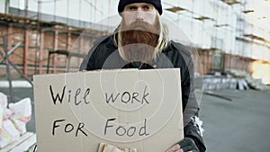 Tilt up of portrait of young homeless man with cardboard looking at camera and wants to work for food standing near