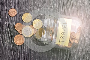 Tilted glass jar with scattered euro coins with the inscription TRAVEL on a black wooden table