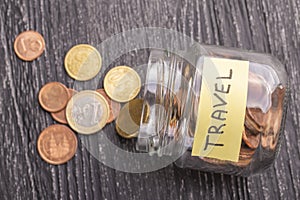 Tilted glass jar with scattered euro coins with the inscription TRAVEL on a black wooden table