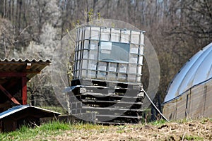 Tilted white intermediate bulk container or IBC plastic tank with metal cage put on top of wooden pallets in local garden to be