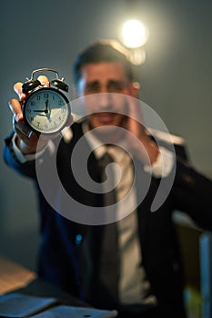 Time is an asset, use it wisely. a stressed out businessman holding a clock during a late night at work.