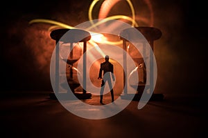 Time concept. Silhouette of a man standing between hourglasses with smoke and lights on a dark background