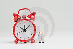 Time counting down for retirement concept, miniature happy senior old couple standing with big red alarm clock on white background