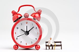 Time counting down for retirement concept, miniature happy senior old man sitting on bench with big red alarm clock on white