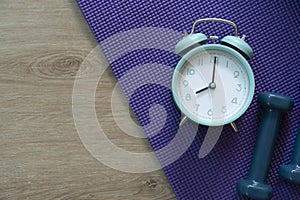 Time for exercising clock and dumbbell with purple yoga mat background, sport and healthy concept