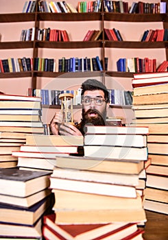 Time flow concept. Man on surprised face holds hourglass while studying, bookshelves on background. Man, scientist in