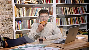 Time lapse caucasian working guy is chilling at workplace, holding pizza slice and drinkink coke while looking at laptop