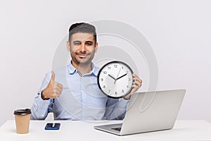 Time management. Cheerful young man employee sitting in office workplace with laptop, holding big clock