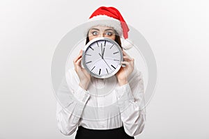 Time management concept - Young business woman with santa hat hiding behind a clock isolated over white background.