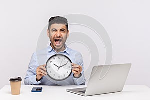 Time management. Furious stressed out man employee sitting at workplace with laptop, holding clock and shouting