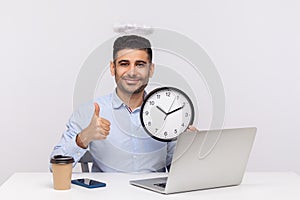 Time management. Joyful punctual man employee with angelic nimbus sitting in office workplace, holding clock