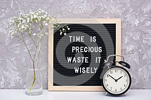 Time is precious waste it wisely. Motivational quote on letterboard, black alarm clock, flower in vase on table against stone wall