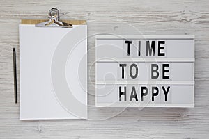 `Time to be happy` words on a modern board, clipboard with blank sheet of paper on a white wooden surface, top view. Overhead,