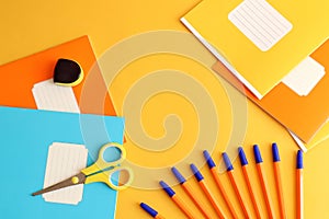 Time to go to school: school notebooks in orange, yellow, blue, sharpener, scissors, pens on a yellow background, top view, space